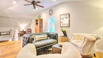A spacious living room with a leather sofa, a white armchair, and a wooden cabinet. A ceiling fan is visible, and there is a window providing natural light.