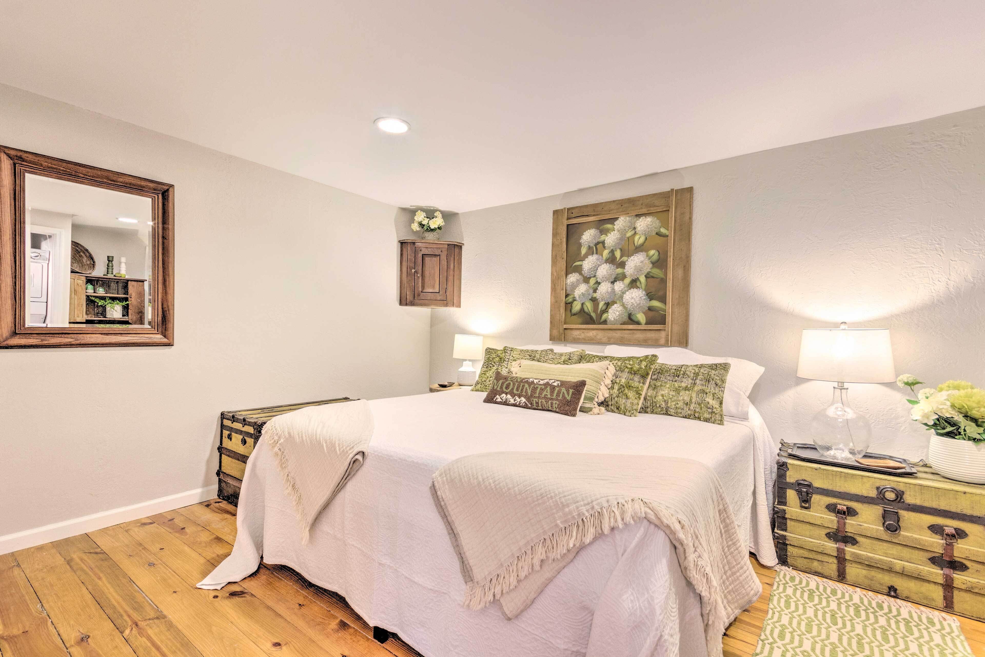 A bedroom with a large bed, wooden floor, a dresser, a mirror, a wall-mounted cabinet, and a painting.