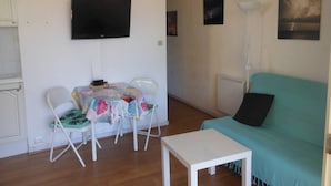 A living room with a teal sofa, a white table, and a small table with chairs. There is a flat-screen TV mounted on the wall.