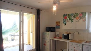 A compact kitchen with a microwave, toaster, and refrigerator, a dining table with a floral tablecloth, and a view through the glass door.