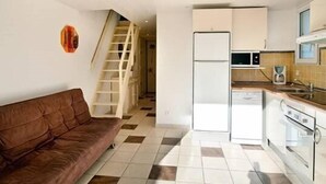 A compact kitchen with a refrigerator, microwave, and stove. There is a brown sofa and a staircase leading to another room. - APT 3 ROOMS MEZ 45m2 + TERRACE 7m2 REF 4007 (MARSEILLAN PLAGE)