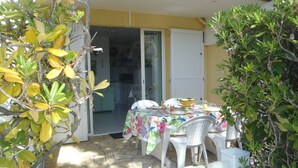 An outdoor patio with a table set for dining, surrounded by greenery and a view of a kitchen through a glass door. - APT 3 ROOMS 35m2 + TERRACE 13m2 REF 4010 (MARSEILLAN PLAGE)
