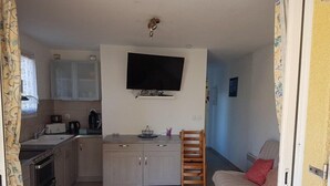 Un espace de vie compact comprenant un coin cuisine, un téléviseur à écran plat fixé au mur, une chaise en bois, une petite table et un canapé agrémenté d’un coussin rouge.