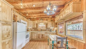 Interior de una cabaña de madera con cocina, que incluye armarios de madera, un refrigerador blanco, una mesa de comedor con sillas y un ventanal con vistas a árboles.