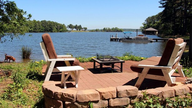 Una terraza junto al lago con dos sillas Adirondack, una mesa pequeña y un muro de piedra.