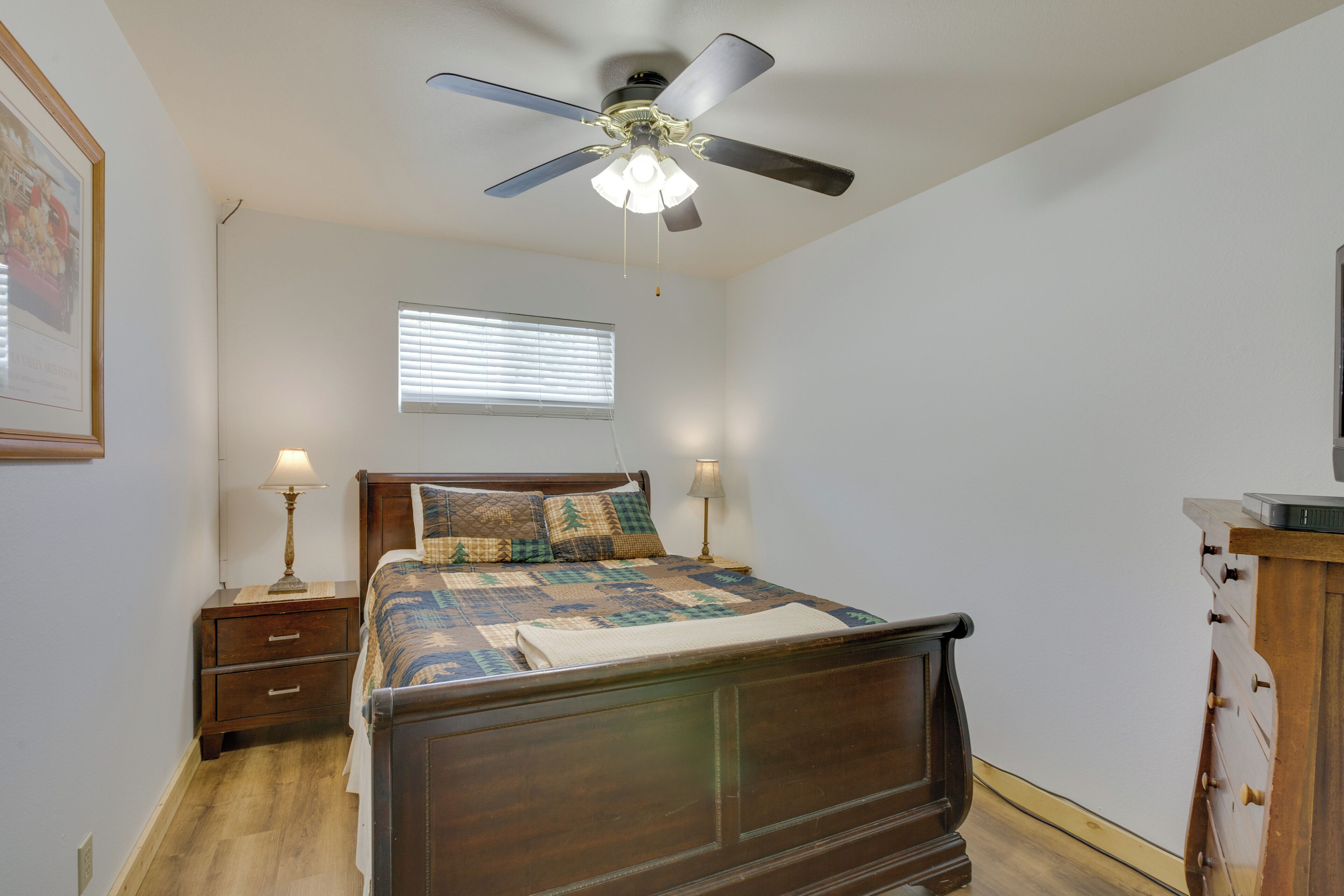 A bedroom with a wooden bed, two bedside tables with lamps, a ceiling fan, a window with blinds, and a framed picture on the wall.