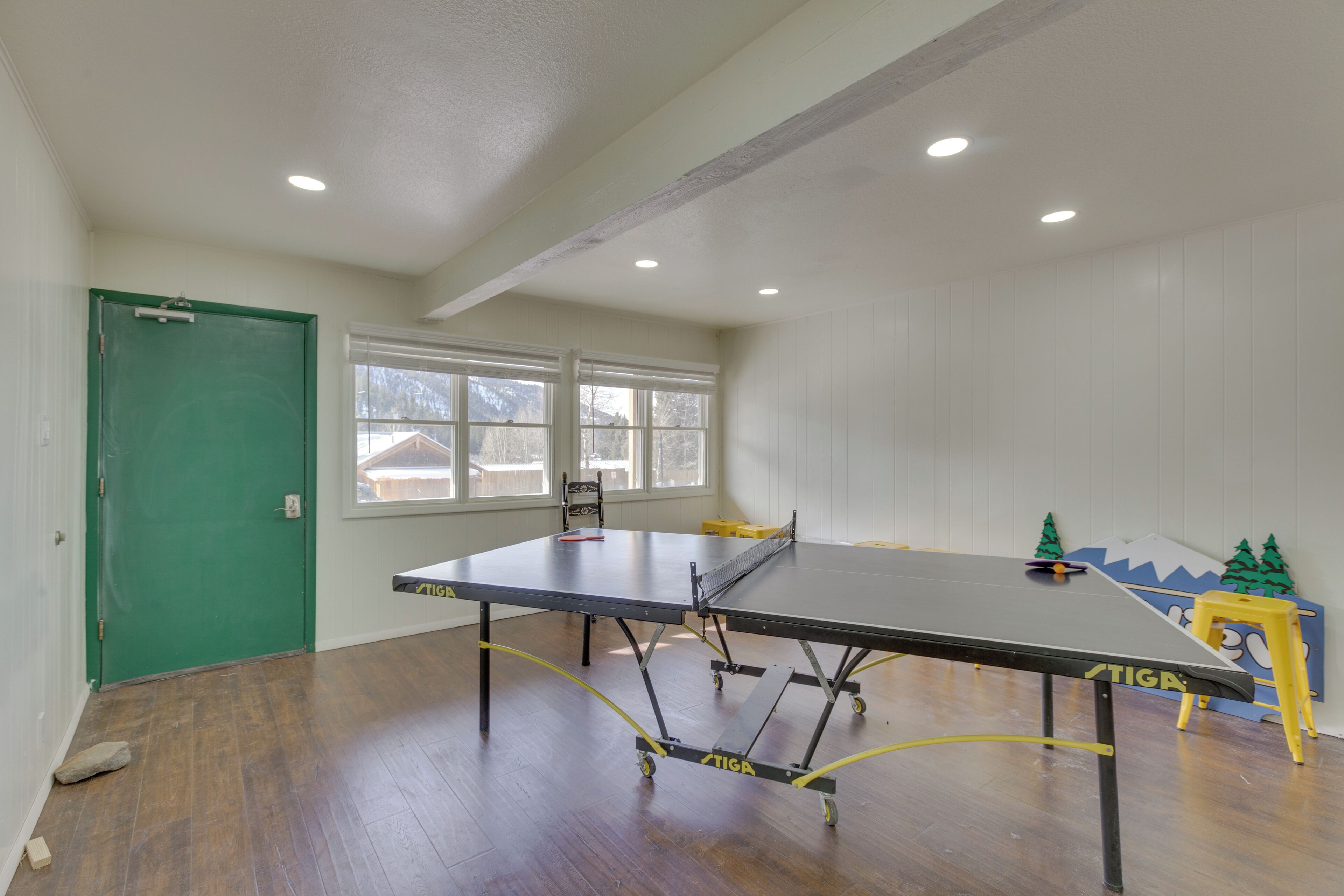 A room with a ping pong table, wooden flooring, a green door, and windows with a view of houses outside.
