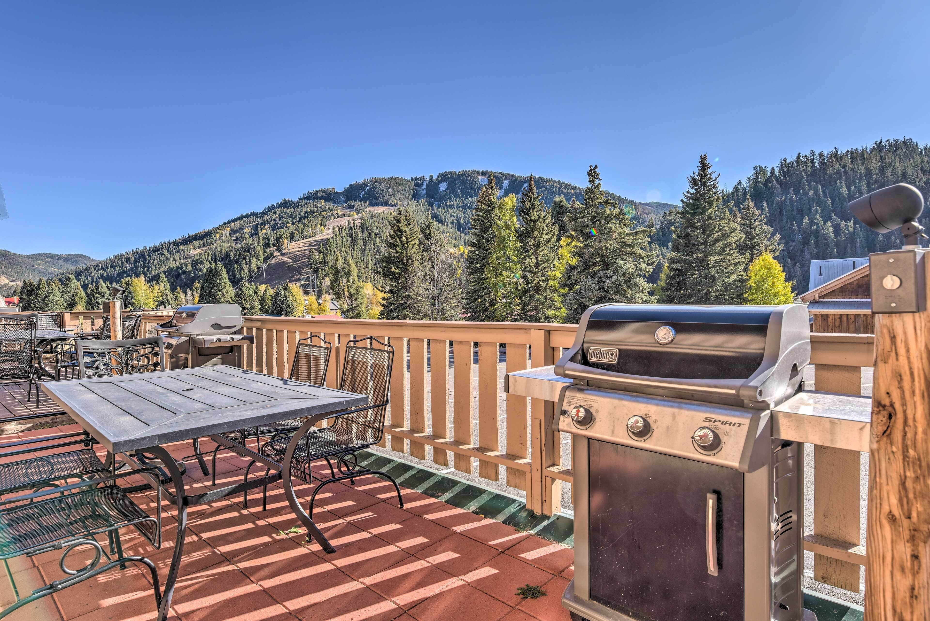 An outdoor patio with a grill, tables, and chairs, overlooking a mountainous landscape.