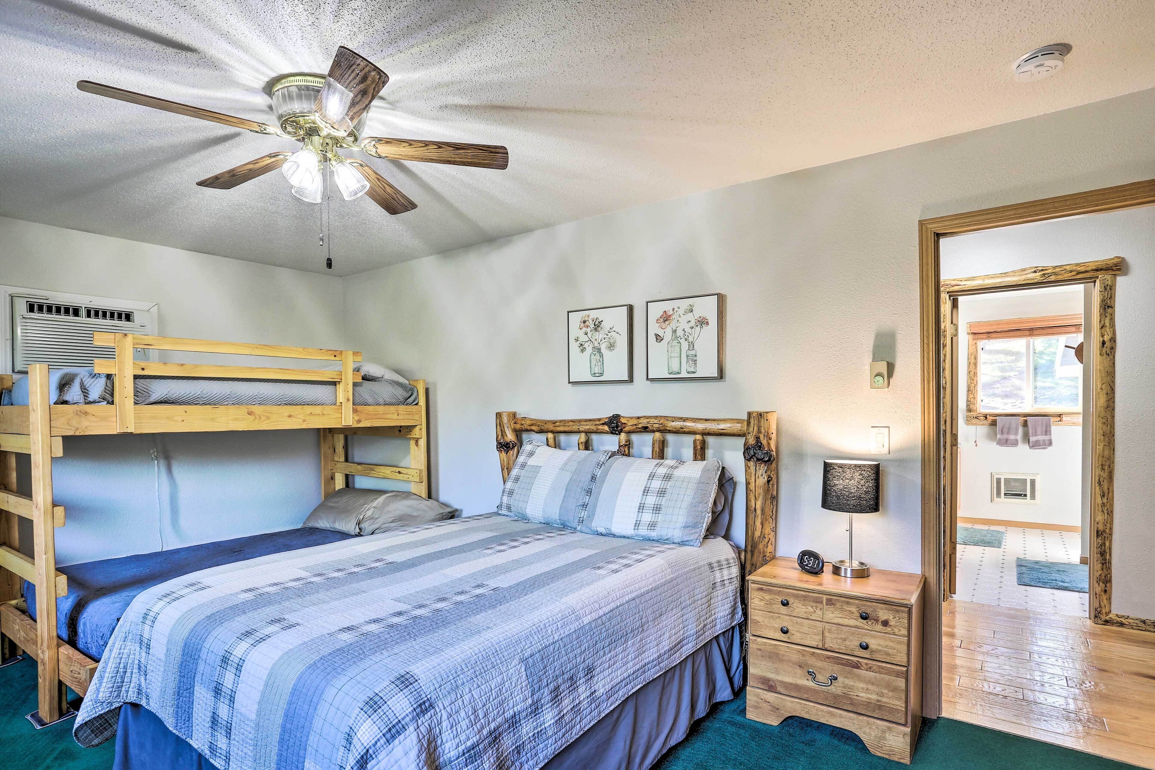 A bedroom with a bunk bed, a single bed with a striped blanket, a wooden headboard, a nightstand with a lamp, and a ceiling fan.