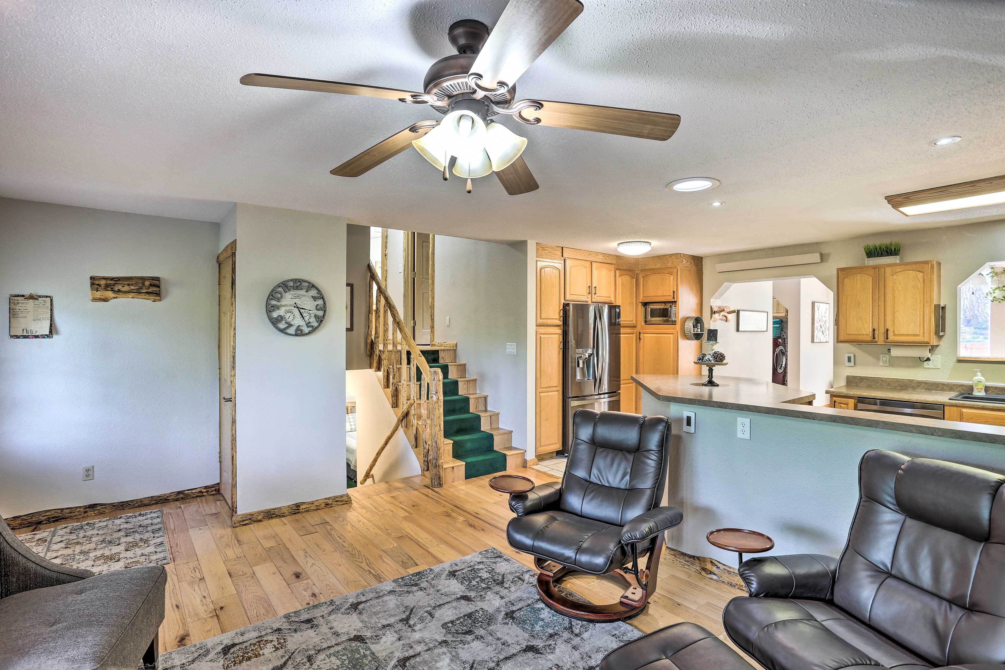 A spacious living room with a ceiling fan, a fireplace, and a view of the kitchen and staircase.