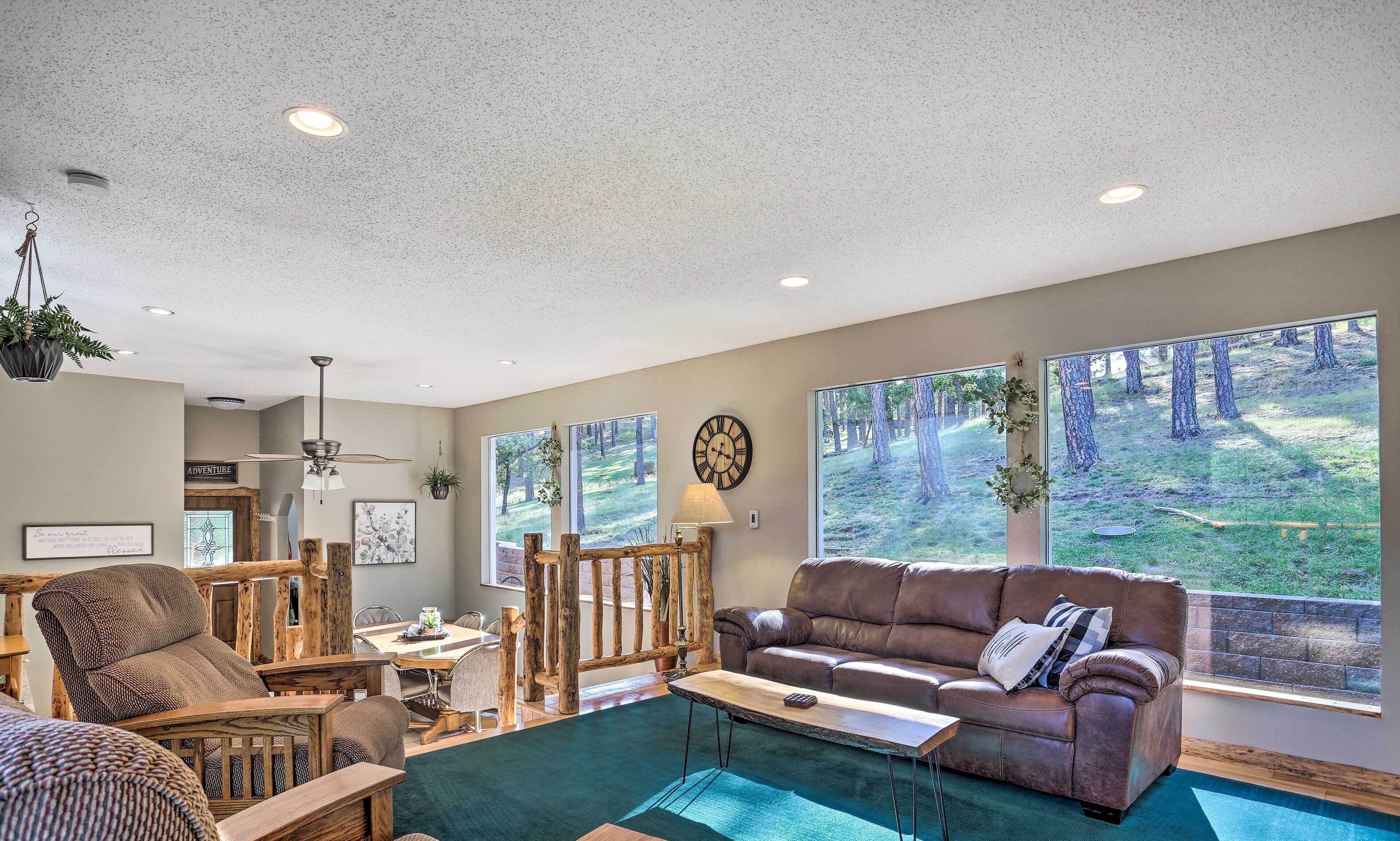 A spacious living room with a large window, a brown leather sofa, a wooden coffee table, and a dining area with a wooden table and chairs.