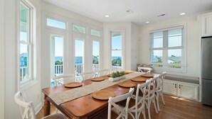 Uma sala de jantar com uma mesa de madeira, cadeiras brancas e vista para o oceano através de grandes janelas.
