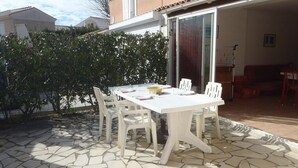 Une terrasse avec une table et des chaises blanches, un sol en pierre et une porte-fenêtre coulissante donnant sur un espace intérieur où se trouve un canapé rouge.