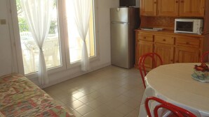 A kitchen with a refrigerator, microwave, and wooden cabinets. There is a dining table with chairs and a clock on the wall. - APT 3 ROOMS 36m2 + LOGGIA  7m2 REF 4031 (MARSEILLAN PLAGE)