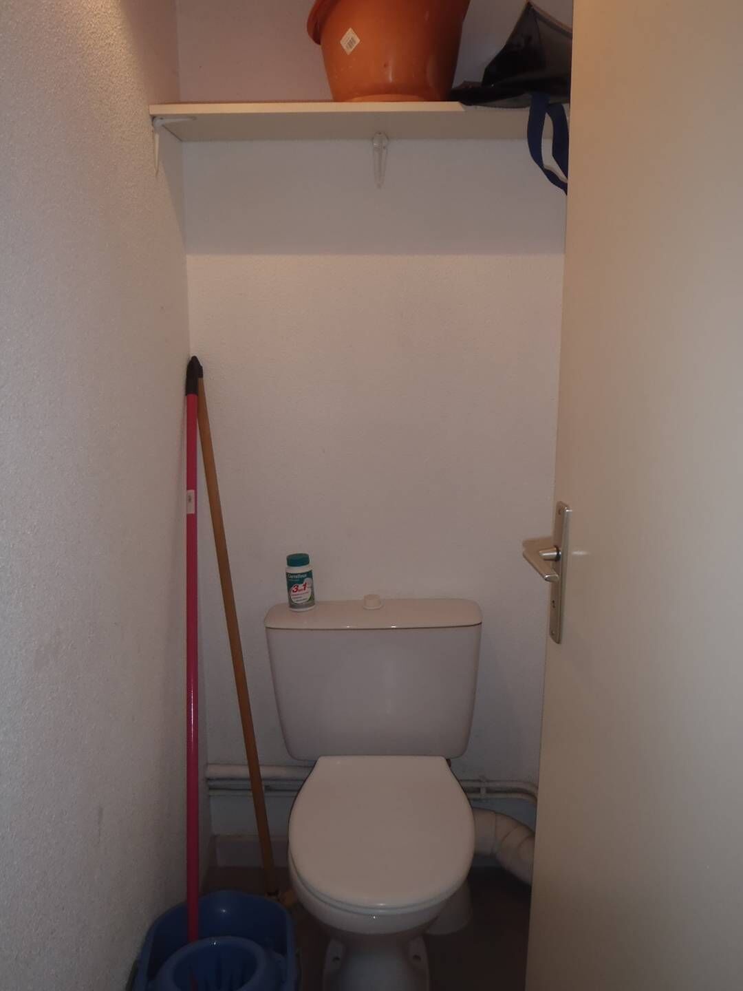 A small, compact bathroom with a toilet, a broom, and a bucket.