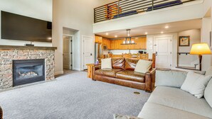 A spacious living area with a fireplace, a leather sofa, a high ceiling, and a kitchen in the background. - Ski-in/ski-out Granby Penthouse w/ Mountain Views! (Granby)