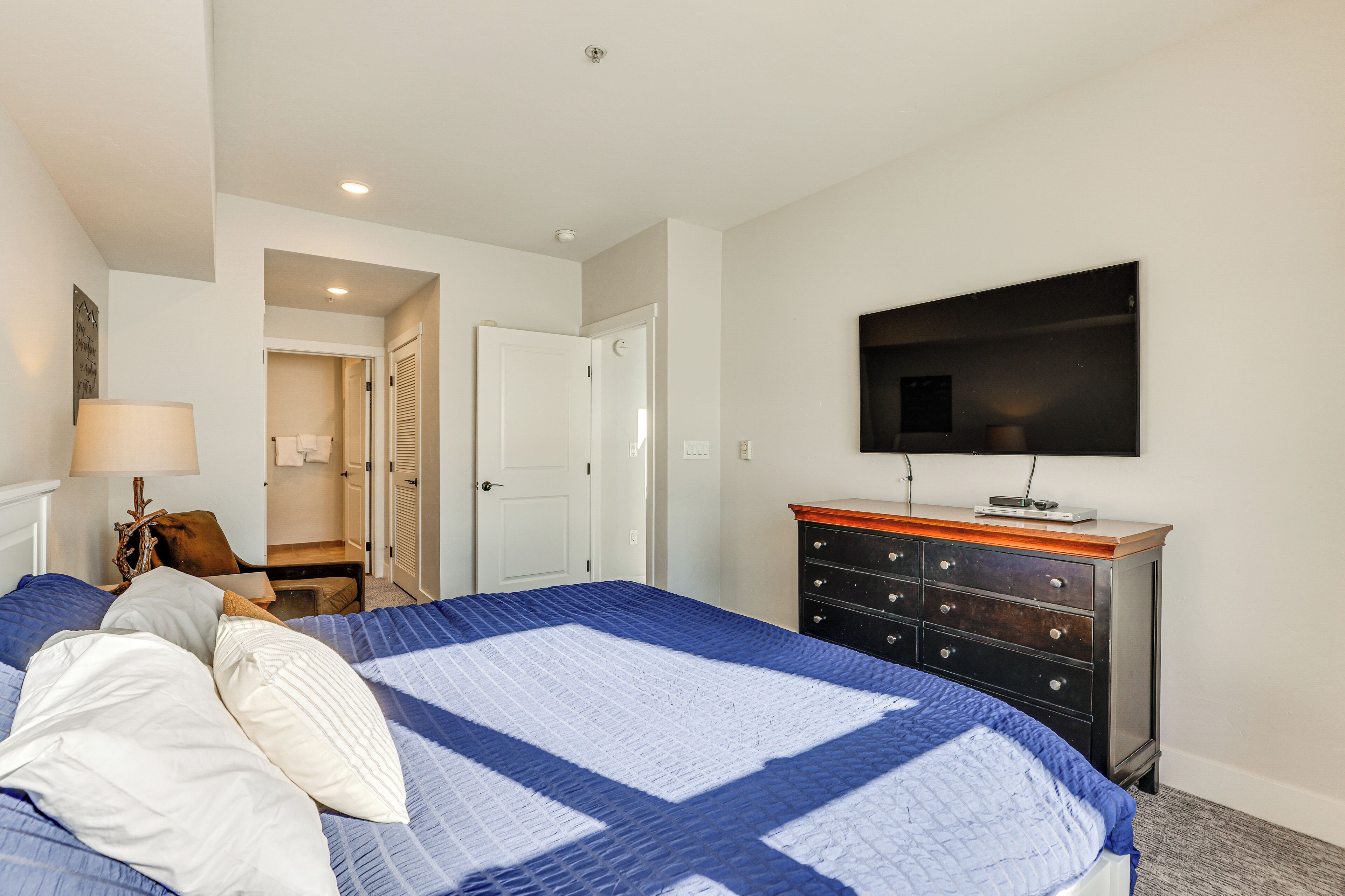 Une chambre à coucher avec un grand lit, une commode, un téléviseur fixé au mur et une porte donnant sur une autre pièce.