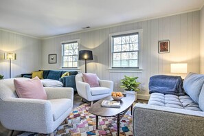 A living room with a sofa, two armchairs, a coffee table, and a floor lamp. There are windows with white frames and a plant on the windowsill. - Charming Carrboro Home - Walk to Downtown! (Carrboro)