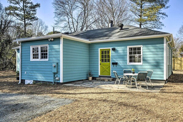 A small, single-story house with a green door and windows, a patio with chairs, and a stone pathway leading to the door. - Charming Carrboro Home - Walk to Downtown! (Carrboro)
