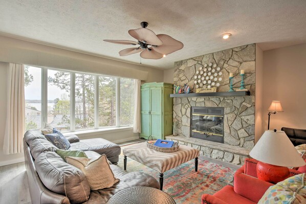 A living room with a stone fireplace, a ceiling fan, and a large window.
