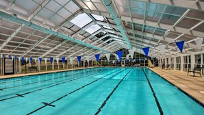 Piscina interna com pistas demarcadas, teto alto com claraboias e bandeiras triangulares azuis e brancas.