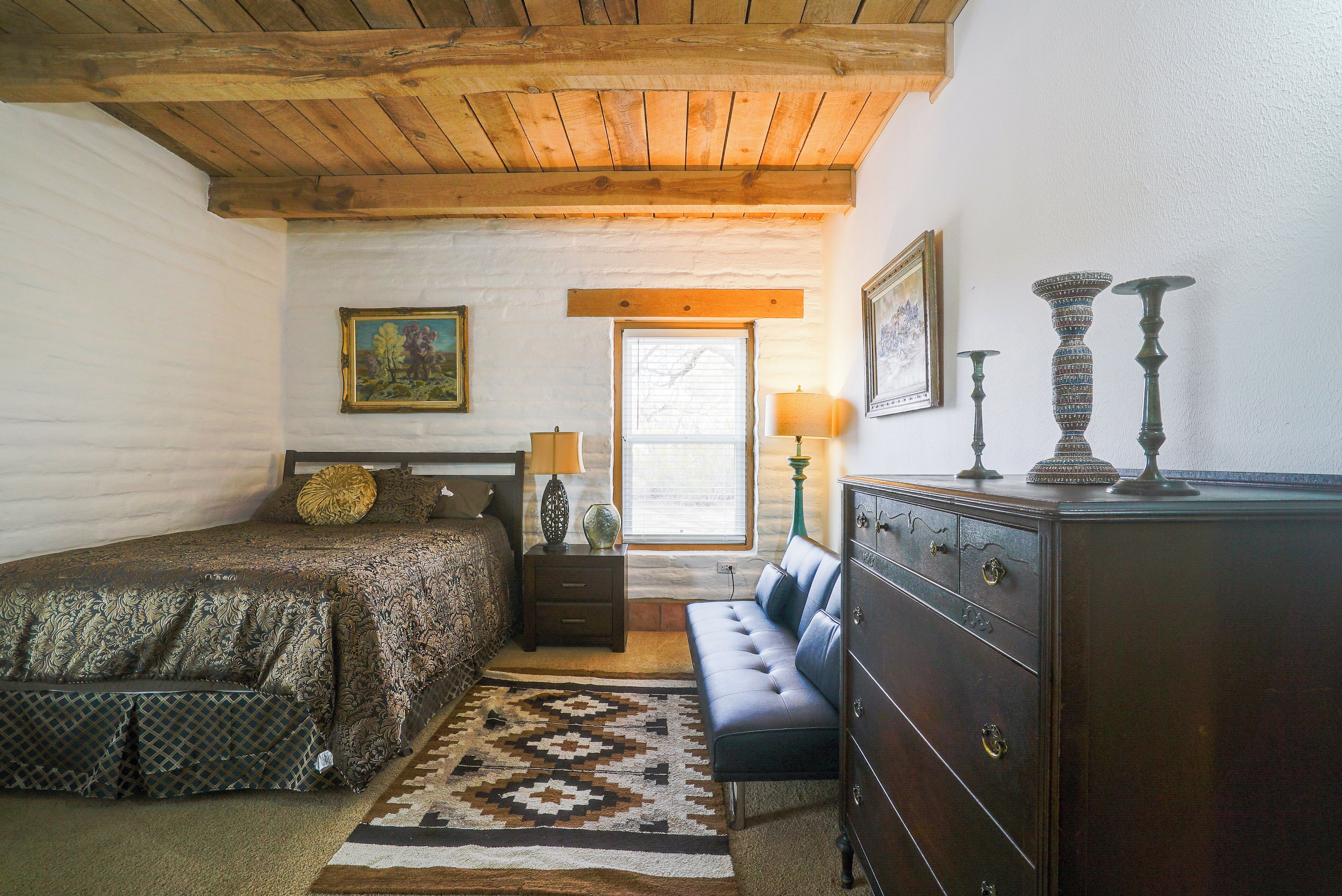 A bedroom with a bed, a dresser, a bench, a rug, and a window with blinds.
