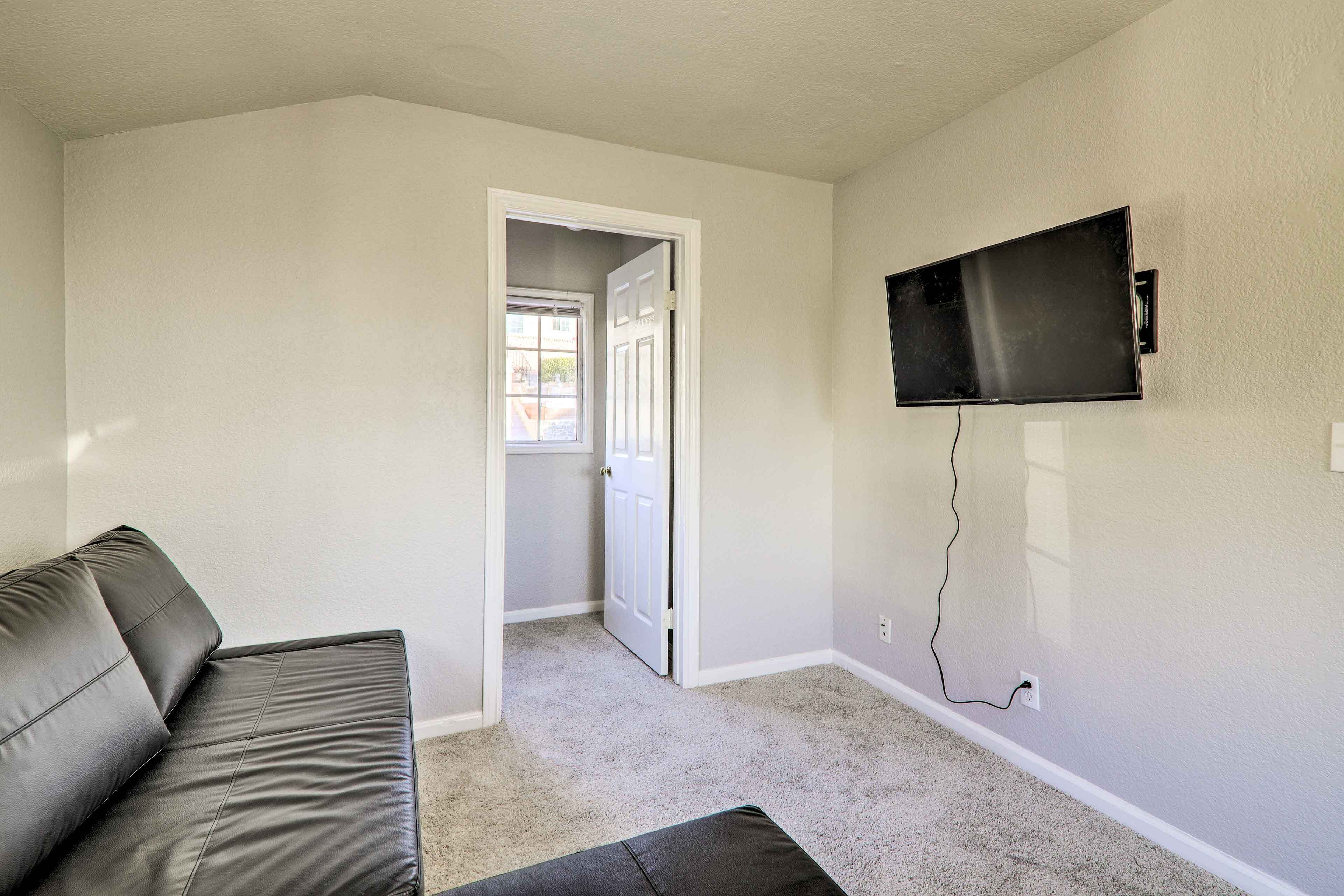 A room with a grey leather sofa, a flat-screen TV mounted on the wall, and a carpeted floor.