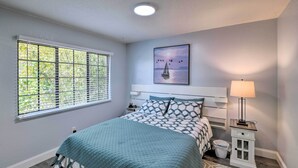 Une chambre à coucher comprenant un lit, une table de chevet, une lampe, une fenêtre avec des stores et un tableau accroché au mur.