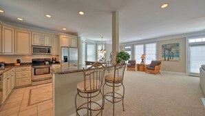 A spacious kitchen with modern appliances, a dining area with two bar stools, and a cozy seating area with a plant and a painting. - Manteo Waterfront Resort Home w/ 30-ft Dock! (Manteo)