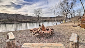A lakeside cabin with a fire pit surrounded by logs and stones, a wooden deck, and a view of the lake and hills. - La Petite Maison in Mount Olive w/ Fire Pit! (Mountain View)