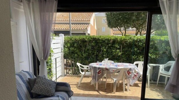 A living area with a sofa, a table, and chairs, a sliding glass door, and a view of a garden. - VILLA  3 ROOMS MEZ 48m2 + TERRACE 7m2 REF 4034 (MARSEILLAN PLAGE)