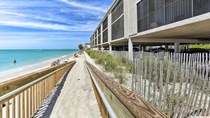 Un paseo marítimo frente a la playa con barandilla de madera, un sendero de arena y un edificio con grandes ventanales.