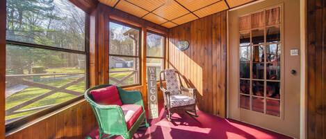A wooden-paneled room with a rocking chair, a chair with a floral cushion, and a large window offering a view of a yard.