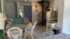 A compact living space with a sofa, two white chairs, a dining table with a plate of fruit, a small kitchen area with a microwave, and a staircase. - VILLA  3 ROOMS MEZ 57m2 + TERRACE 11m2 REF 4036 (MARSEILLAN PLAGE)
