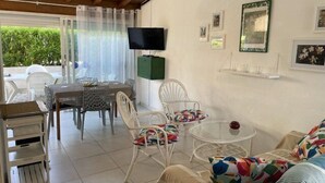A furnished room with a dining area, a TV, and a view of greenery outside. - VILLA  3 ROOMS MEZ 57m2 + TERRACE 11m2 REF 4036 (MARSEILLAN PLAGE)