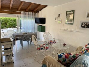 A furnished room with a dining area, a TV, and a view of greenery outside.