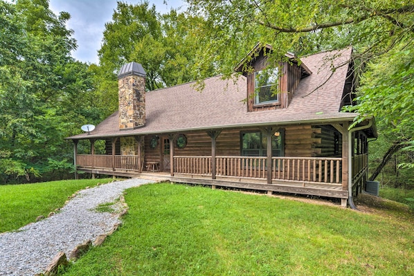 A log cabin with a stone chimney, a porch with railings, and a gravel path leading to the entrance.
