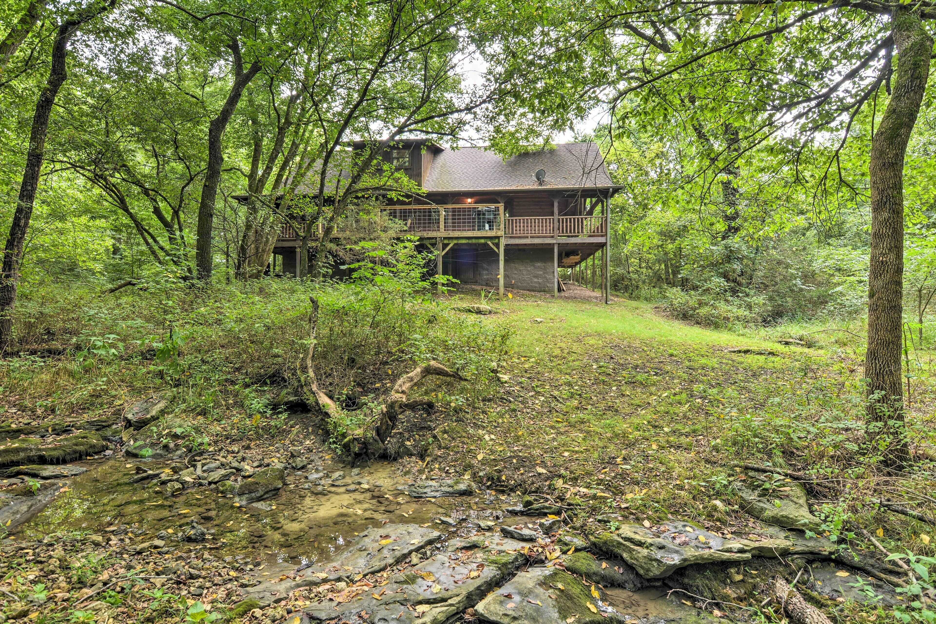 A cabin with a deck, surrounded by dense forest and a stream.