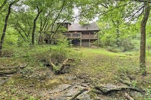 A cabin with a deck, surrounded by dense forest and a stream.