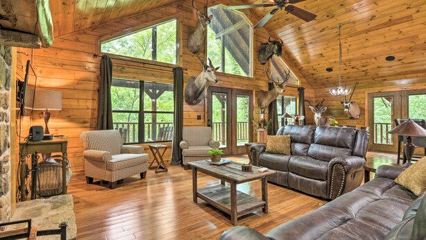 A rustic cabin interior with wooden walls, a ceiling fan, leather sofas, and mounted animal heads.