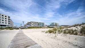 Ein hölzerner Steg führt zu Strandhäusern mit mehreren Balkonen und einem klaren Himmel.