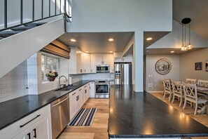 A modern kitchen with white cabinets, a black countertop, stainless steel appliances, and a dining area with wooden chairs. - Lake Pend Oreille Home w/ Dock & Paddle Boards (Sagle)