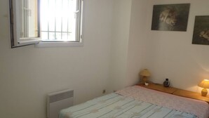 A bedroom with a bed, a window, a radiator, a nightstand with a lamp, and two wall-mounted pictures. - APT 3 ROOMS 32m2 + TERRACE 13m2 REF 4050 (MARSEILLAN PLAGE)
