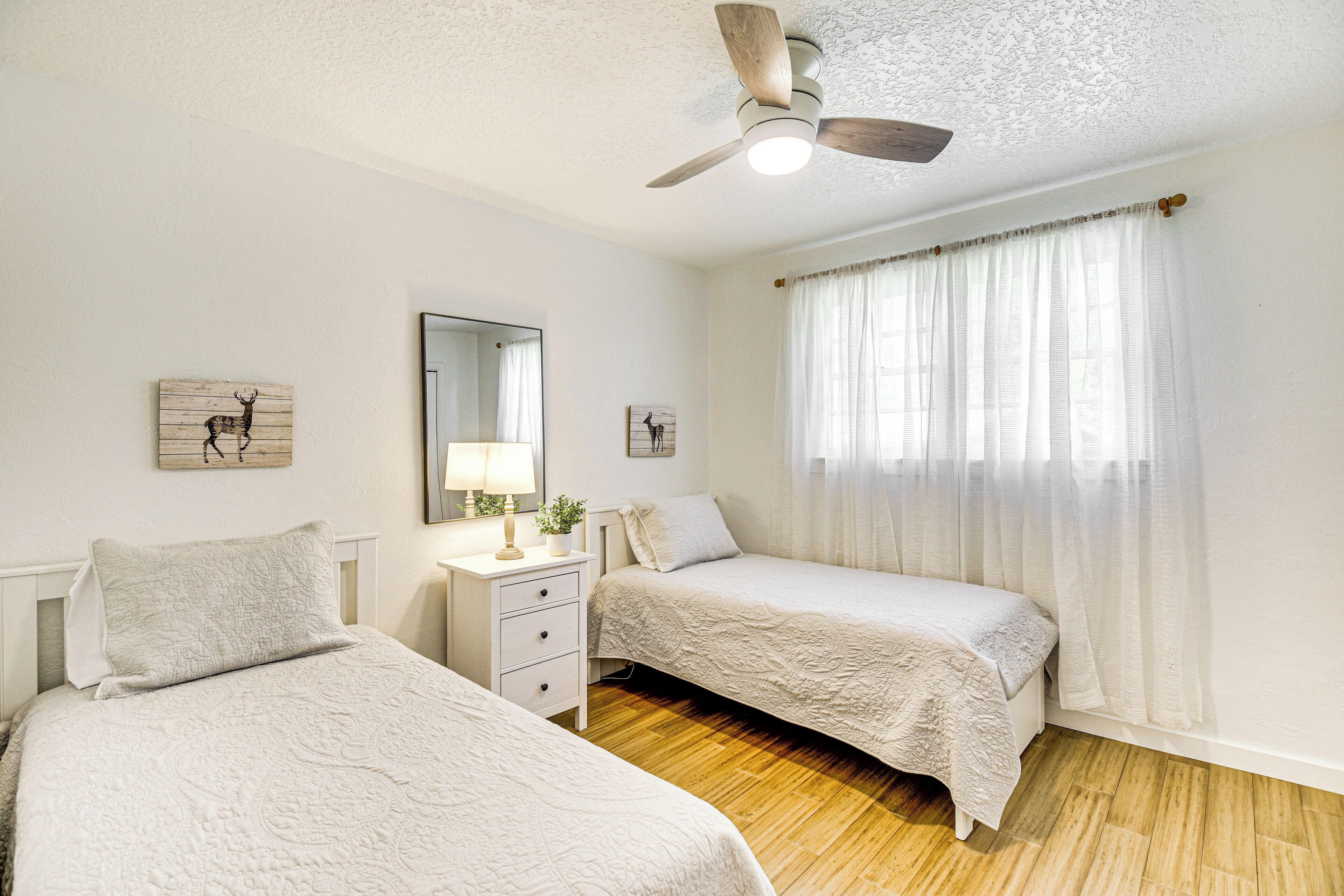 A bedroom with two beds, a ceiling fan, a nightstand with a lamp, and a window with sheer curtains.