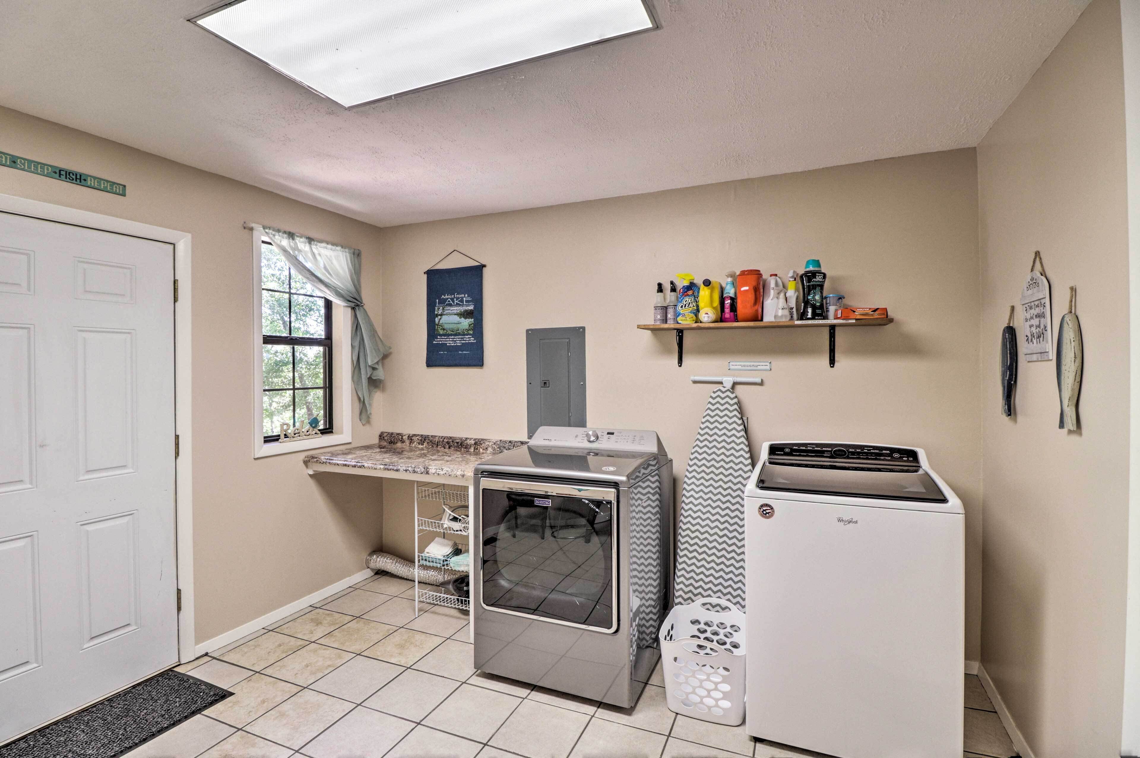 A laundry room with a dryer, washing machine, and ironing board. There is a shelf with cleaning supplies and a window with curtains.