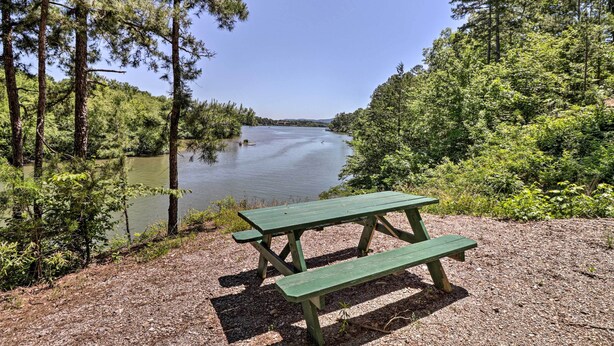 Une table de pique-nique verte, avec des bancs, entourée d’arbres et bordée par un plan d’eau.