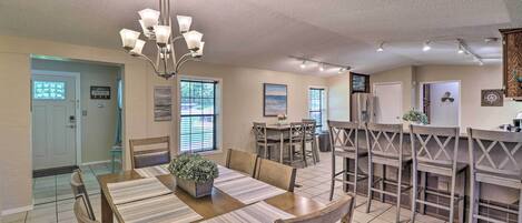 A spacious dining area with a wooden table and chairs, a chandelier, and a kitchen area with bar stools.