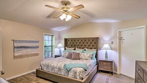 Une chambre à coucher avec un grand lit, un ventilateur de plafond, deux tables de chevet avec des lampes, une porte et une fenêtre avec des stores.