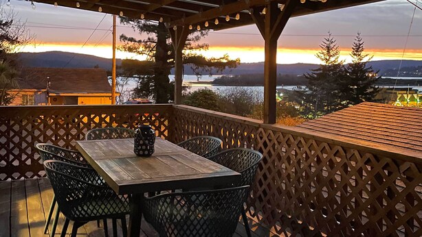 A wooden deck with a table and chairs, overlooking a body of water and distant mountains during sunset.