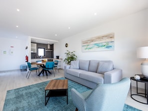 A modern living room with a grey sofa, a wooden coffee table, and a blue patterned armchair. A kitchen with white cabinets and a window is visible in the background. - Villa Drop In with private pool and garden in Sagres (Sagres)